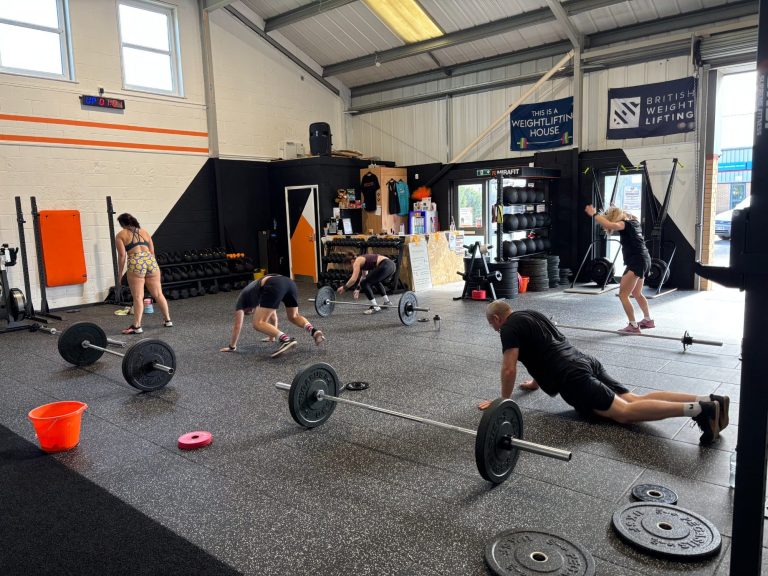 Two men in workout gear, one assisting the other on the gym floor near a barbell.